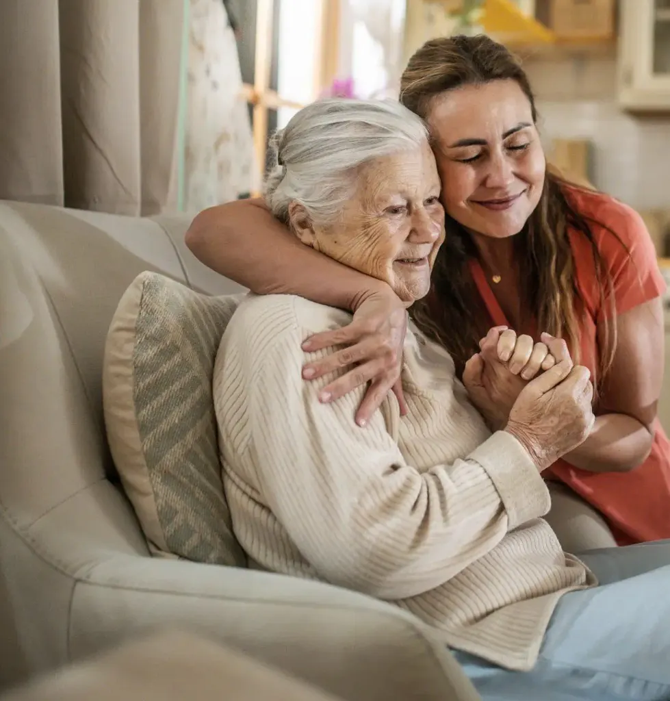 A carer sitting with an older woman, arm around her shoulders.